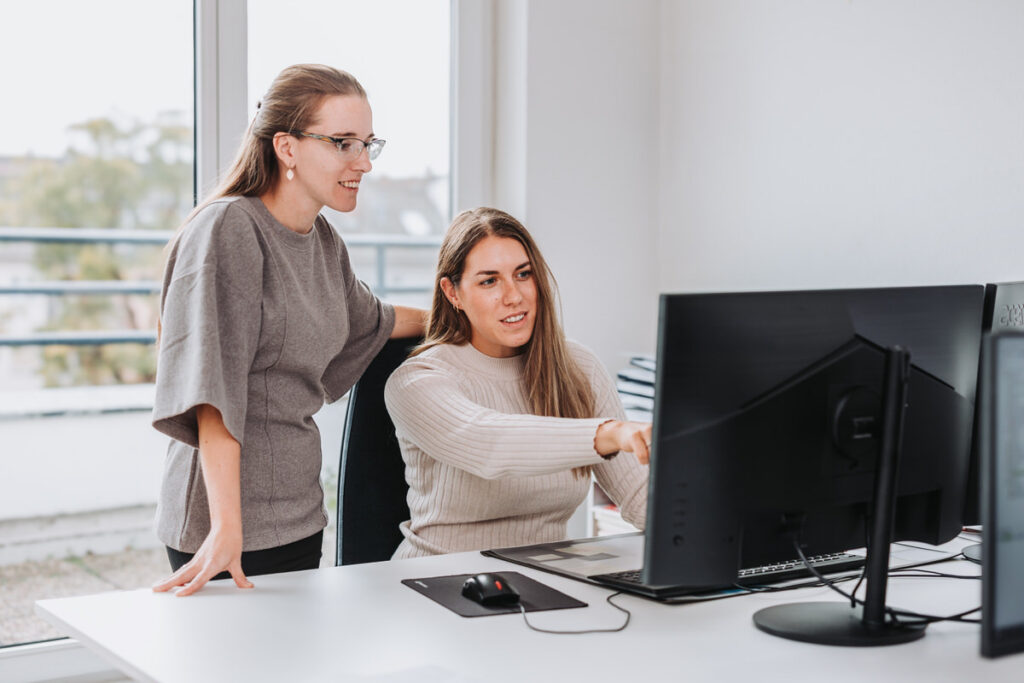 Zwei Frauen unterhalten sich in einem hellem Raum. Die Frau mit einem beigen Shirt sitzt vor dem Monitor und erklärt der Person neben sich stehend etwas am Bildschirm.