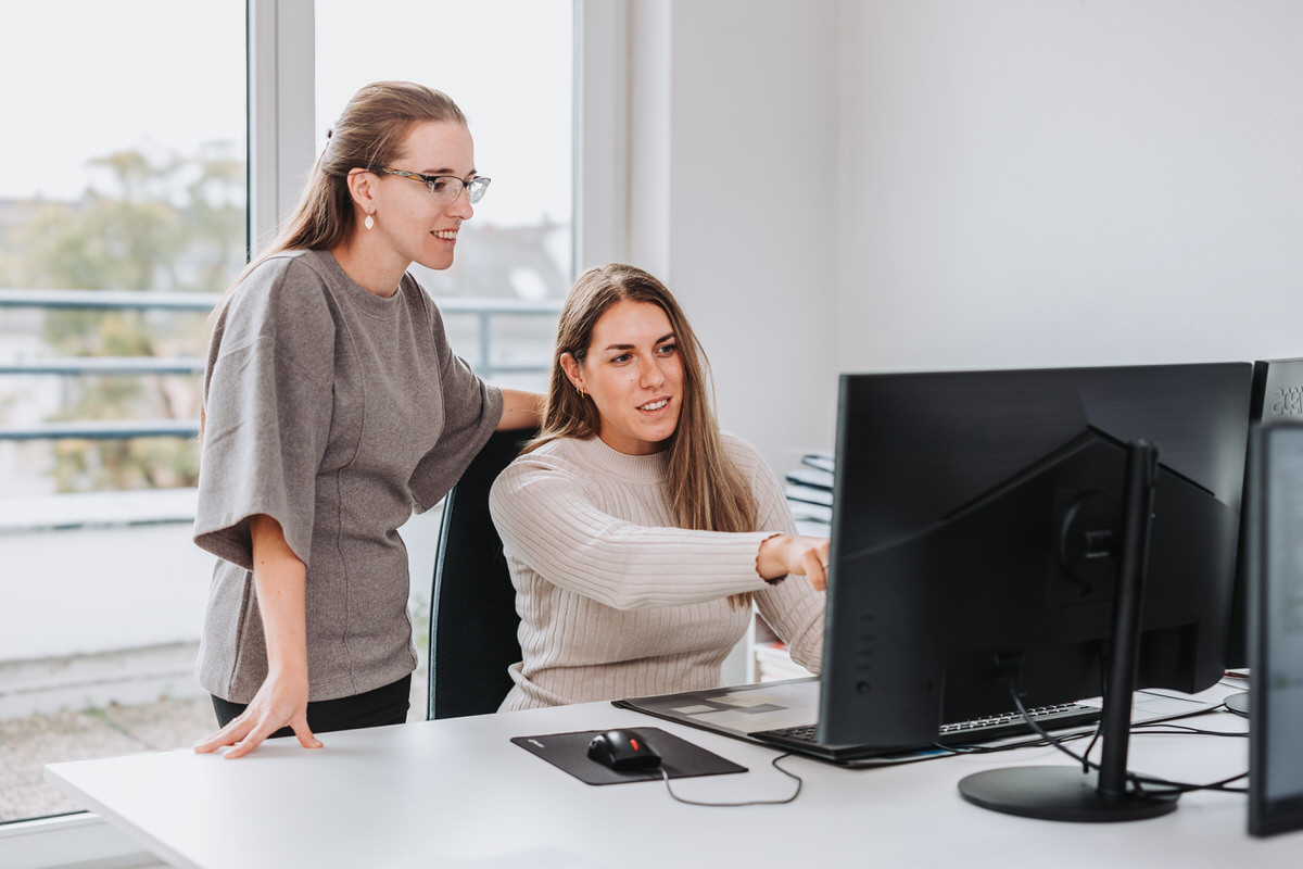 Zwei Frauen unterhalten sich in einem hellem Raum. Die Frau mit einem beigen Shirt sitzt vor dem Monitor und erklärt der Person neben sich stehend etwas am Bildschirm.